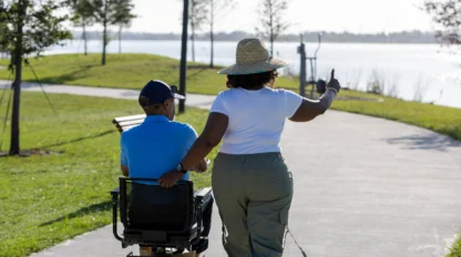 A woman in a wide-brimmed hat walks beside a person in a mobility scooter on a lakeside path, gesturing toward the water.