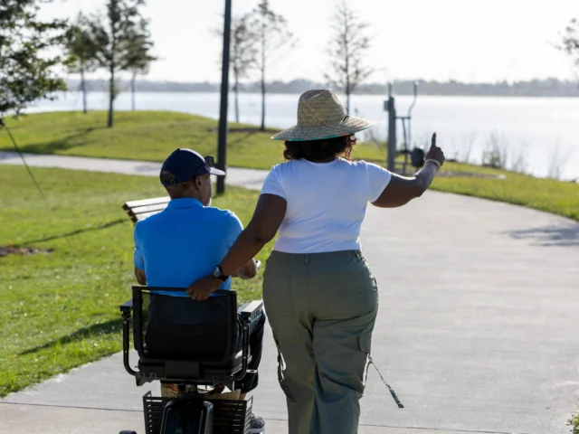 A woman in a wide-brimmed hat walks beside a person in a mobility scooter on a lakeside path, gesturing toward the water.