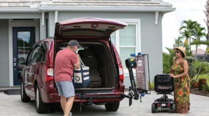 A man in a red van loads items into the back using a lift. A woman stands nearby with a mobility scooter, outside a suburban house.