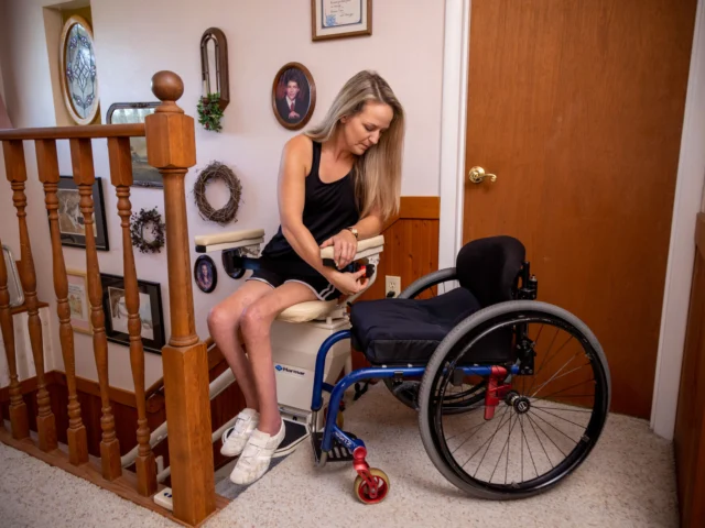 A woman in a stairlifts is transferring from a stairlift seat to a wheelchair in a hallway adorned with family photos and decorations.