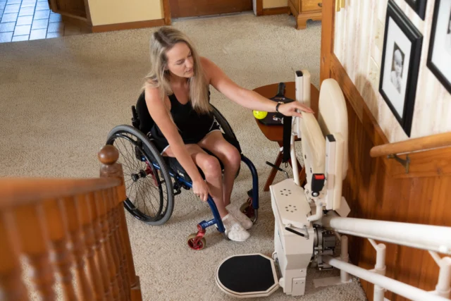 A woman in a wheelchair is adjusting the footrest of a stair lift in a carpeted hallway with framed photos on the wall.