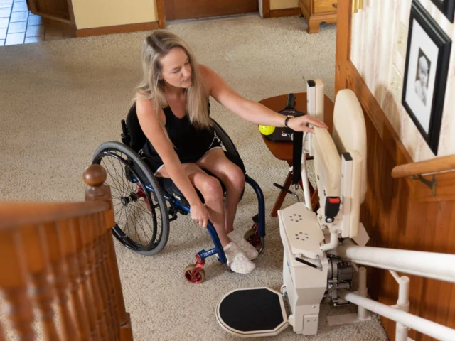 A woman in a wheelchair is adjusting the footrest of a stair lift in a carpeted hallway with framed photos on the wall.