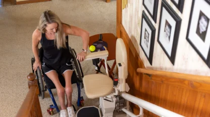 A woman transitions from a wheelchair to a stairlift at the base of a carpeted staircase. Framed photos are visible on the wall nearby.