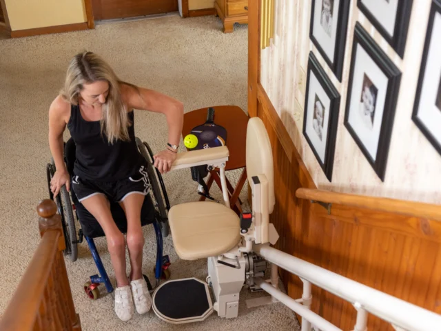 A woman transitions from a wheelchair to a stairlift at the base of a carpeted staircase. Framed photos are visible on the wall nearby.