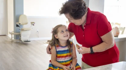 Elderly woman in a red shirt smiles at a young girl in a colorful dress, who is seated and smiling back, in a bright room with a chair lift visible in the background.
