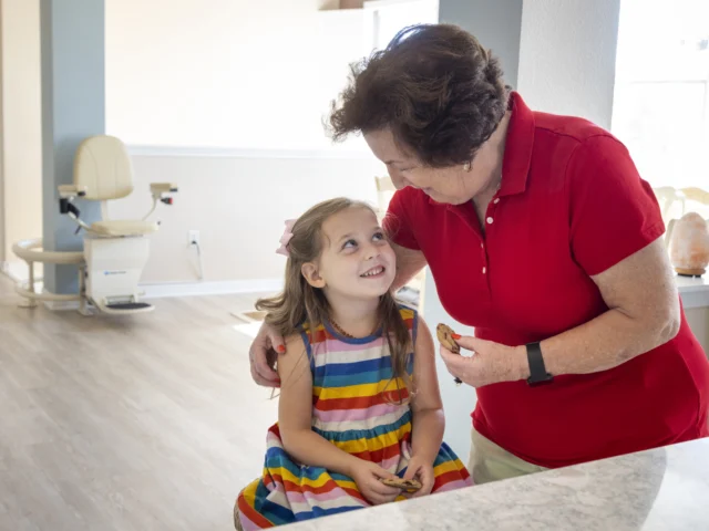 Elderly woman in a red shirt smiles at a young girl in a colorful dress, who is seated and smiling back, in a bright room with a chair lift visible in the background.