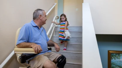 Man seated on a stairlift looks at a young girl in a colorful dress standing on the stairs. They are indoors, next to a handrail.