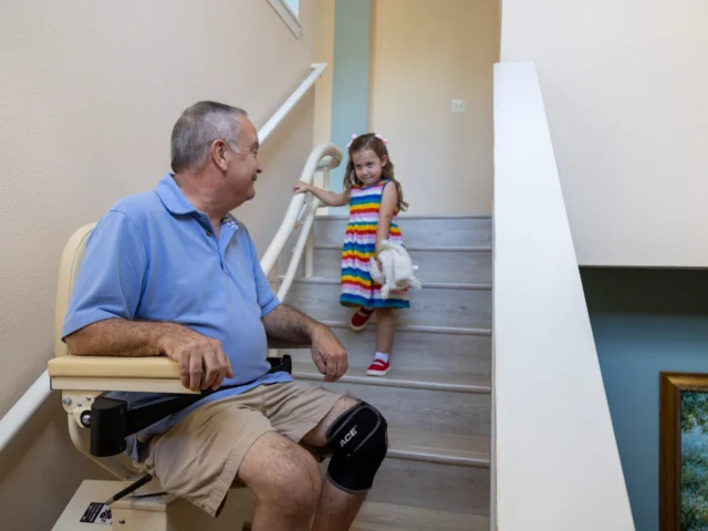 Man seated on a stairlift looks at a young girl in a colorful dress standing on the stairs. They are indoors, next to a handrail.