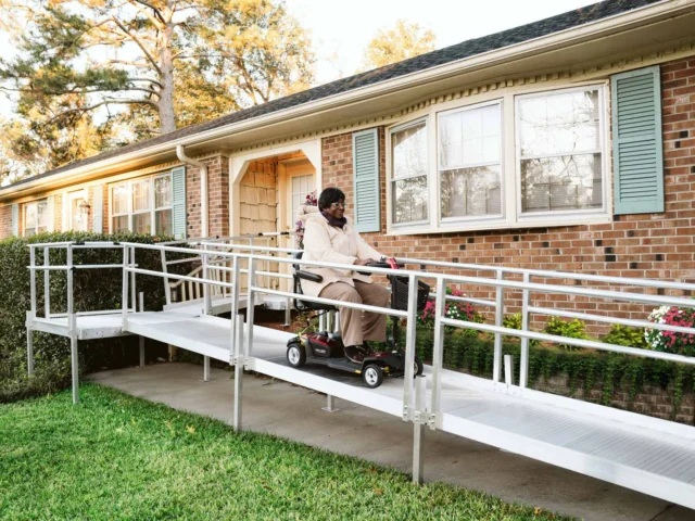 Person using a mobility scooter ascends a residential ramp beside a brick house with green shutters.