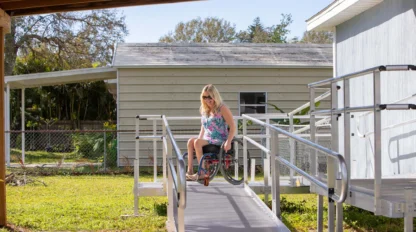 A woman in a wheelchair descends a ramp outside a house, under a covered area. Trees and a fence are visible in the background.