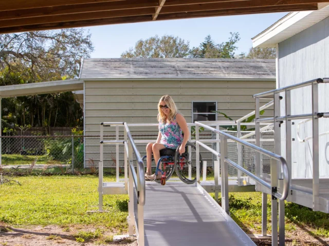 A woman in a wheelchair descends a ramp outside a house, under a covered area. Trees and a fence are visible in the background.