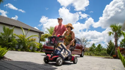 Older adult couple outdoors, with the woman in a mobility scooter carrying a tote bag. They are on a paved path, surrounded by tropical plants and cloudy skies.