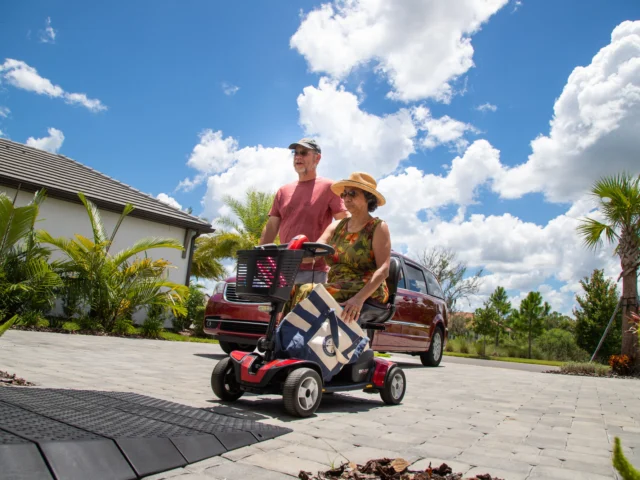 Older adult couple outdoors, with the woman in a mobility scooter carrying a tote bag. They are on a paved path, surrounded by tropical plants and cloudy skies.