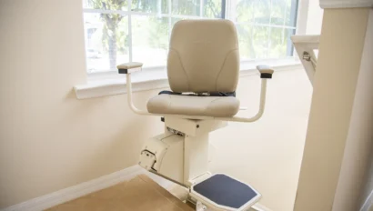 A stairlift chair is positioned at the bottom of a staircase next to a window, with armrests and a footrest.