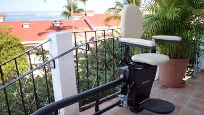 A stairlift installed on an outdoor staircase with a decorative railing, set against a backdrop of red rooftops, trees, and a view of a distant body of water.