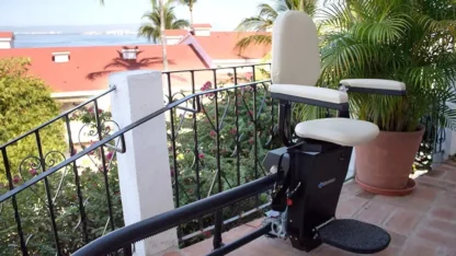 A stairlift installed on an outdoor staircase with a decorative railing, set against a backdrop of red rooftops, trees, and a view of a distant body of water.
