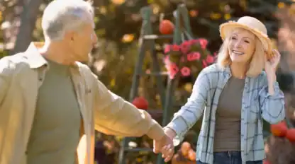 An older couple holds hands outdoors in a garden, with a woman smiling and adjusting her hat. Flowers and pumpkins are visible in the background.