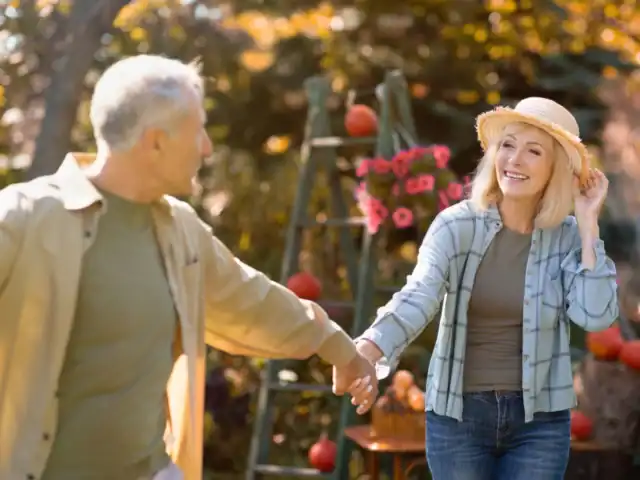 An older couple holds hands outdoors in a garden, with a woman smiling and adjusting her hat. Flowers and pumpkins are visible in the background.