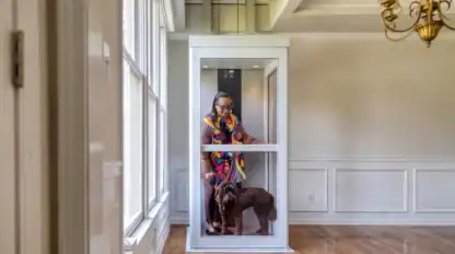 A person wearing colorful clothing stands inside a small home elevator with a brown dog in a bright room with wood floors and white walls.