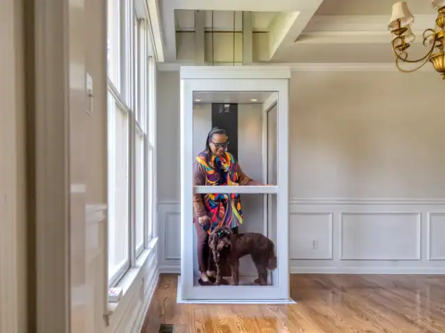 A person wearing colorful clothing stands inside a small home elevator with a brown dog in a bright room with wood floors and white walls.