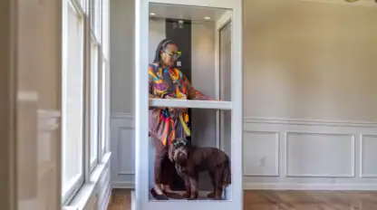 An older woman and a brown dog stand together inside a small residential elevator in a brightly lit room with wood floors and white walls.