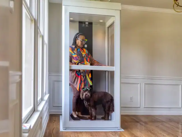 An older woman and a brown dog stand together inside a small residential elevator in a brightly lit room with wood floors and white walls.