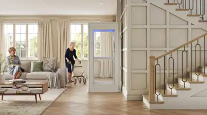 Two older women are in a living room; one sits on a sofa while the other uses a walker to exit a small home elevator near a staircase.