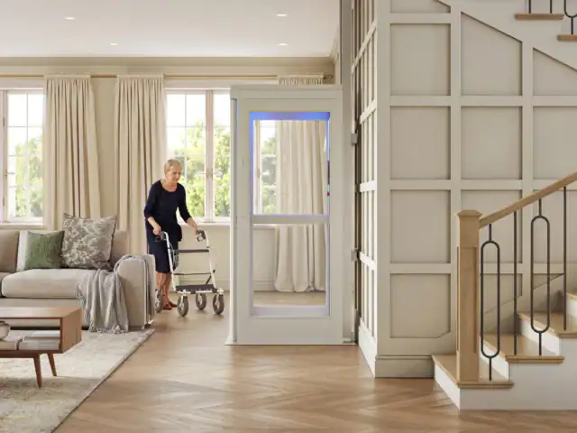 Two older women are in a living room; one sits on a sofa while the other uses a walker to exit a small home elevator near a staircase.