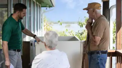 A man in a green shirt explains equipment to an elderly man and woman on a porch, with trees and water visible in the background.