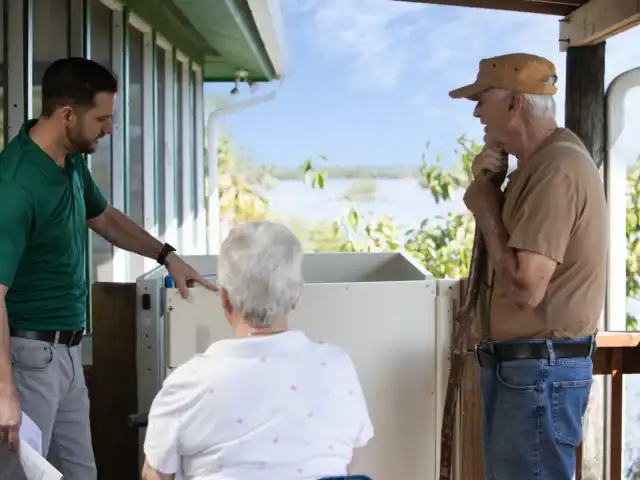 A man in a green shirt explains equipment to an elderly man and woman on a porch, with trees and water visible in the background.