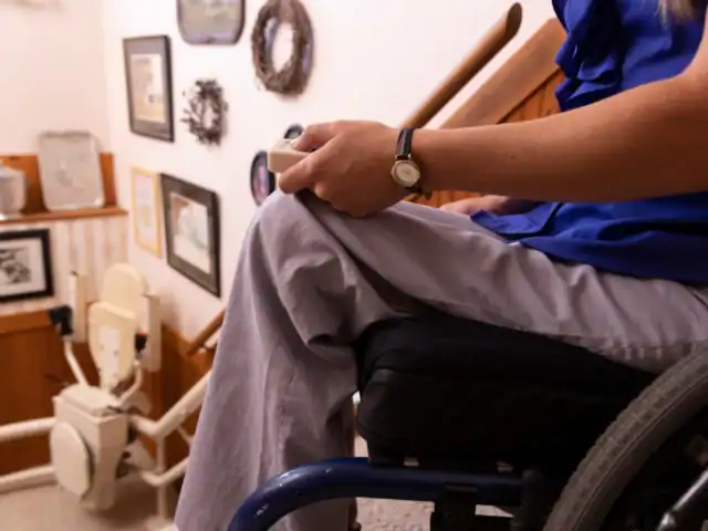 A person in a wheelchair operates a stairlift remote control at the bottom of a staircase decorated with framed pictures and wreaths.
