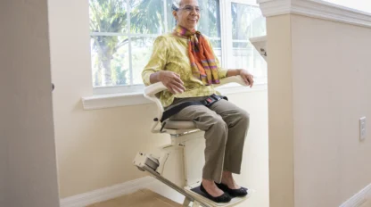 An older woman rides a stairlift in a bright, modern home, wearing glasses, slacks, a yellow blouse, and a colorful scarf.
