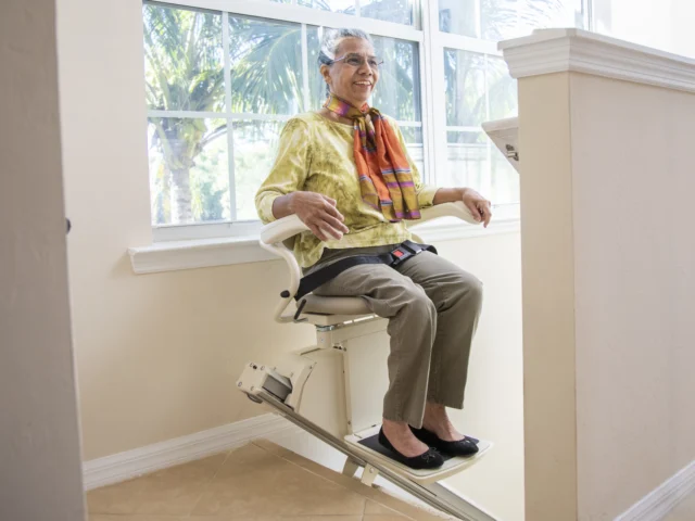 An older woman rides a stairlift in a bright, modern home, wearing glasses, slacks, a yellow blouse, and a colorful scarf.