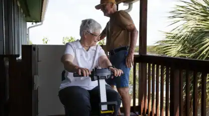 An elderly woman sits on a platform lift on a porch while an elderly man stands nearby, holding the porch railing.