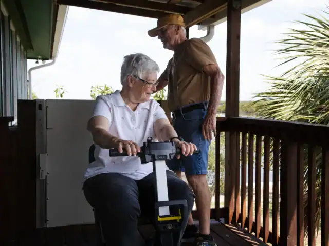 An elderly woman sits on a platform lift on a porch while an elderly man stands nearby, holding the porch railing.