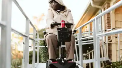 A person wearing a hooded coat uses a mobility scooter on an outdoor ramp next to a building.