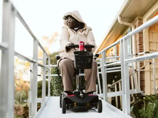 A person wearing a hooded coat uses a mobility scooter on an outdoor ramp next to a building.