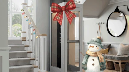 A small residential elevator with a red plaid bow stands near a staircase; holiday lights are on the railing and a decorative snowman is nearby in a modern entryway.