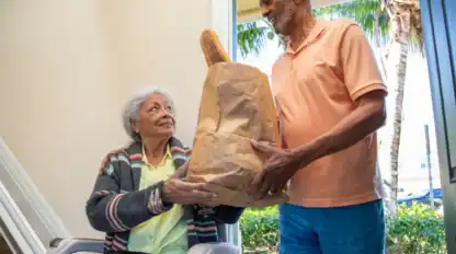 An elderly man standing at a doorway hands a grocery bag with a baguette to an elderly woman sitting in a wheelchair inside the house.