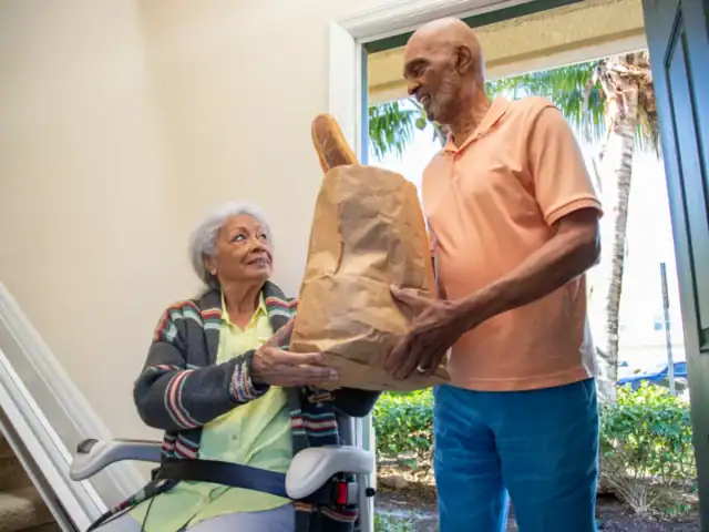 An elderly man standing at a doorway hands a grocery bag with a baguette to an elderly woman sitting in a wheelchair inside the house.