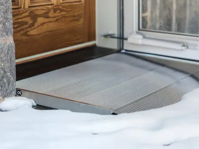 A metal wheelchair ramp leads up to a wooden door, with snow covering the ground outside.