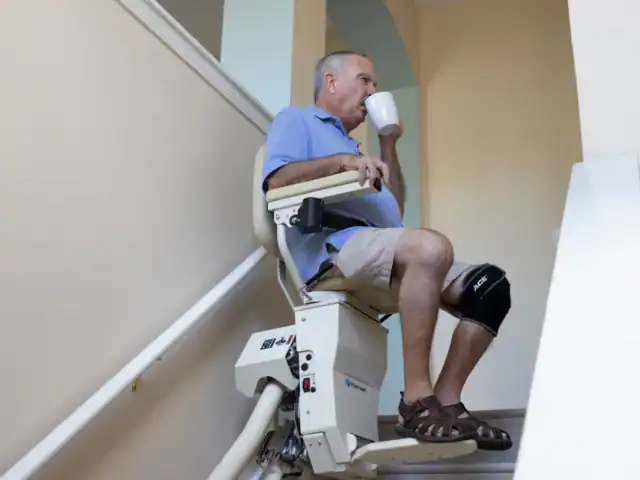An older man with a knee brace drinks from a mug while riding a stair lift up a staircase inside a house.
