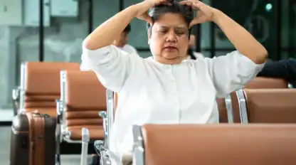 A woman sits in an airport waiting area with her eyes closed and hands on her head, appearing stressed or tired.