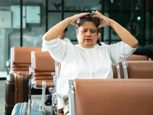 A woman sits in an airport waiting area with her eyes closed and hands on her head, appearing stressed or tired.