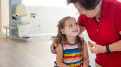 An older woman in a red shirt hugs a young girl in a rainbow-striped dress; both are smiling and holding cookies in a bright indoor setting.