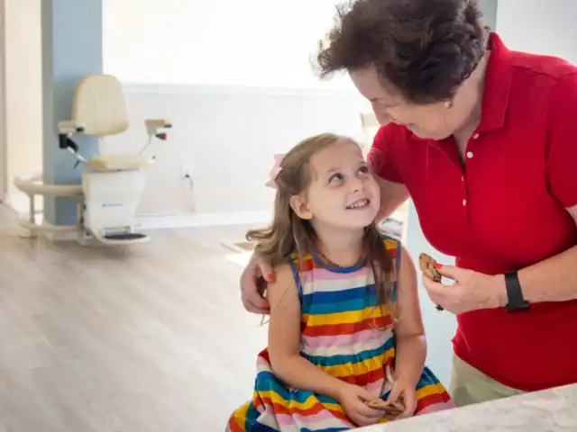 An older woman in a red shirt hugs a young girl in a rainbow-striped dress; both are smiling and holding cookies in a bright indoor setting.