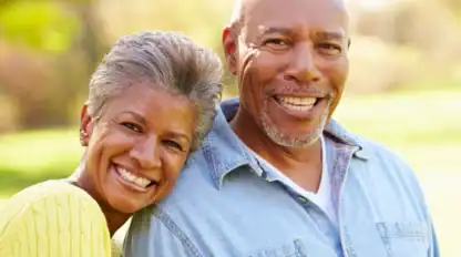 An older woman in a yellow sweater leans her head on the shoulder of an older man in a light denim shirt. Both are smiling outdoors in natural light.