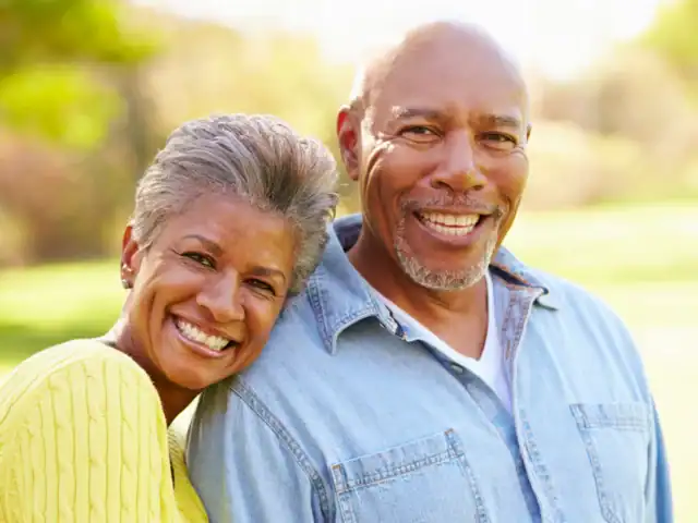 An older woman in a yellow sweater leans her head on the shoulder of an older man in a light denim shirt. Both are smiling outdoors in natural light.
