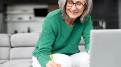 A woman with gray hair and glasses, wearing a green sweater, sits on a couch writing in a notebook while looking at a laptop.