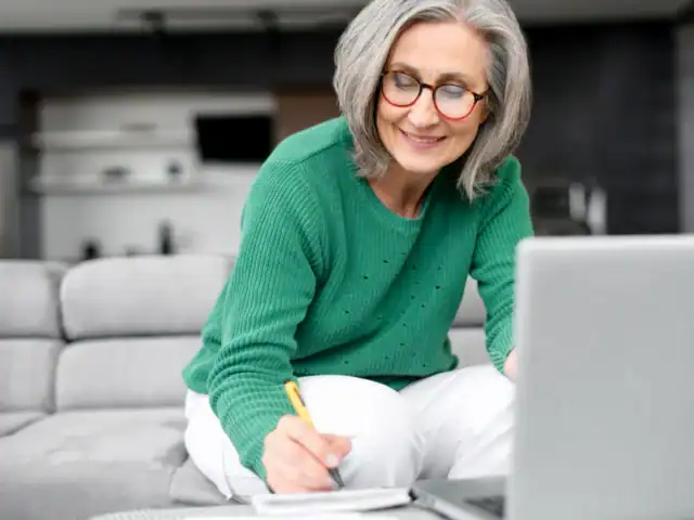 A woman with gray hair and glasses, wearing a green sweater, sits on a couch writing in a notebook while looking at a laptop.